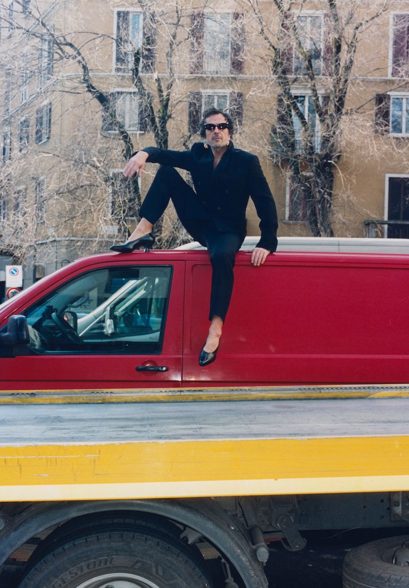 Man wearing a black suit, sunglasses, and heels sitting on top of a red vehicle on a flatbed truck in an urban setting.