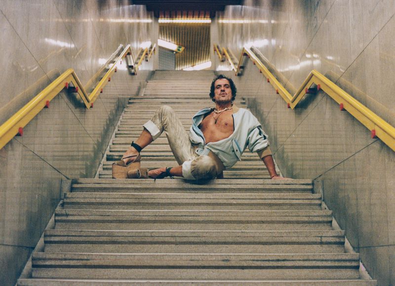 Andreas Kronthaler poses in high platform wedges and an open jacket on a symmetrical subway staircase with yellow handrails.