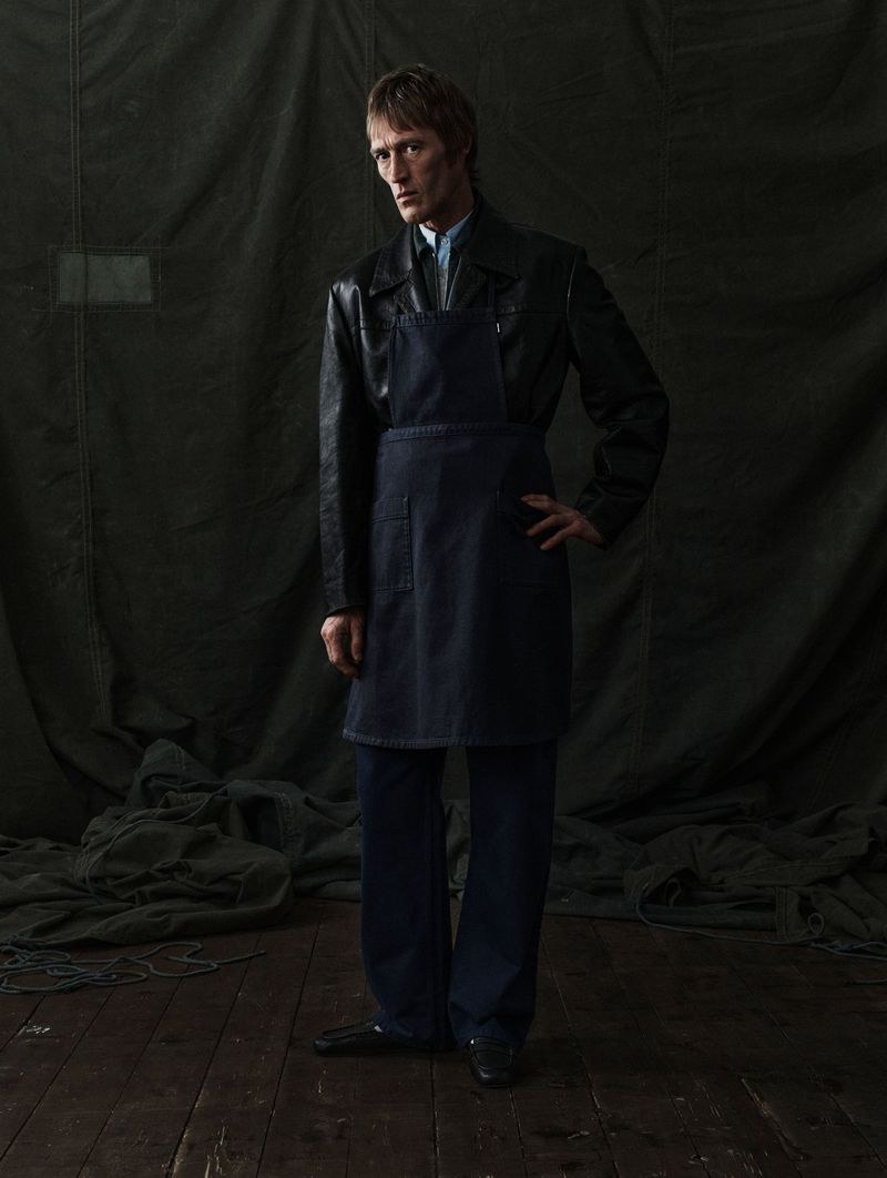 Model Thomas Westcott wearing a navy apron over a black leather jacket against a dark draped canvas backdrop.