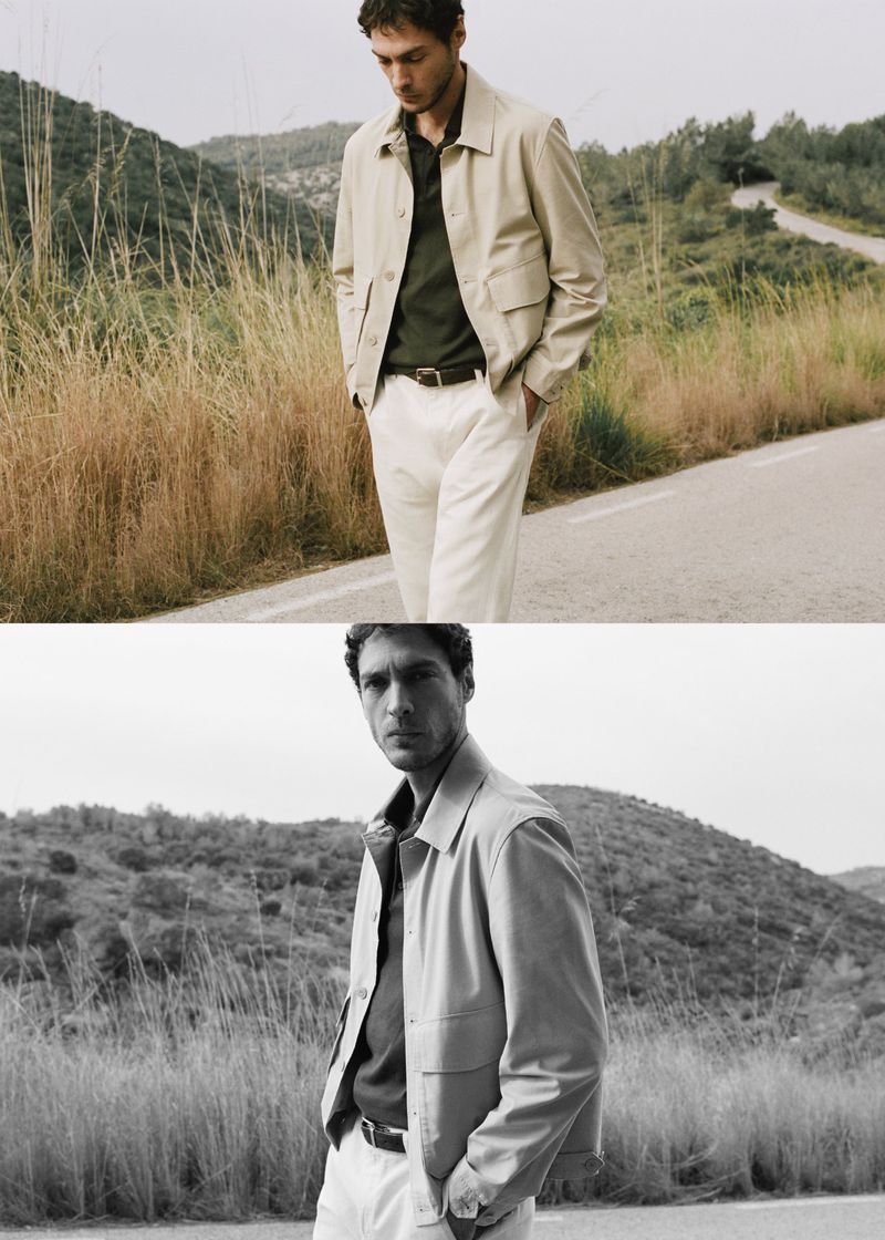 Man wearing a beige jacket and light trousers walking along a country road beside grassy hills under an overcast sky.