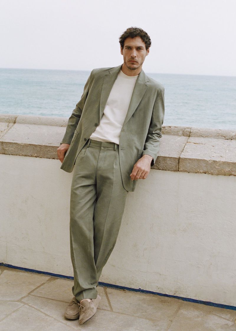 Model Pierre-Benoit Talbourdet in a relaxed sage green tailored suit leaning against a stone balustrade overlooking the ocean.
