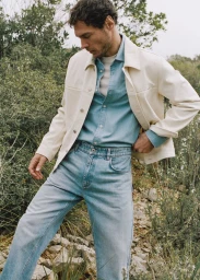 Model Pierre-Benoit Talbourdet wearing an off-white jacket and light blue denim jeans in a natural outdoor landscape.