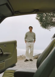 A man in a sage green sweater and cream trousers stands outside near a rocky coast, viewed from inside a vintage car.