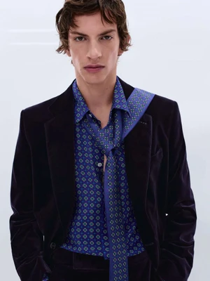 Model wearing a dark velvet blazer and a patterned blue shirt with a matching draped scarf against a white studio background.
