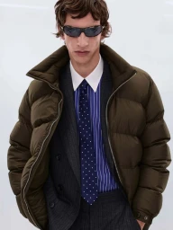 Male model wearing sporty sunglasses, an olive puffer jacket, pinstripe blazer, and polka-dot tie against a white seamless backdrop.