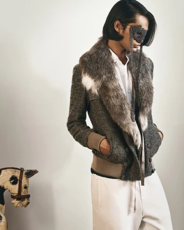 Model in a tweed bomber jacket with a large fur collar and lace eye mask standing against a white studio wall beside a toy horse.