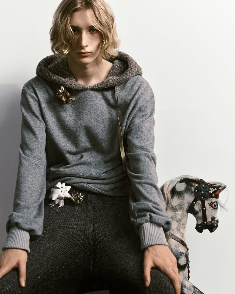 A young blonde man wearing a grey hooded sweatshirt with bow accents sits beside a vintage rocking horse against a white backdrop.