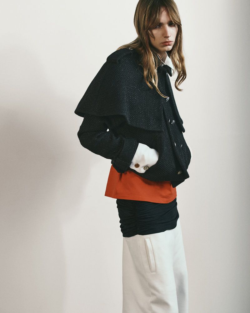 Model wearing a black capelet jacket, rust orange underlayer, and white trousers for Oscar Ouyang AW26 against a white backdrop.