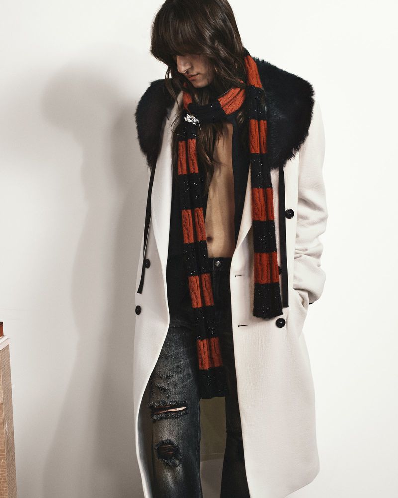 Fashion model wearing a long cream coat with black fur collar, striped knit scarf, and distressed jeans against a white background.