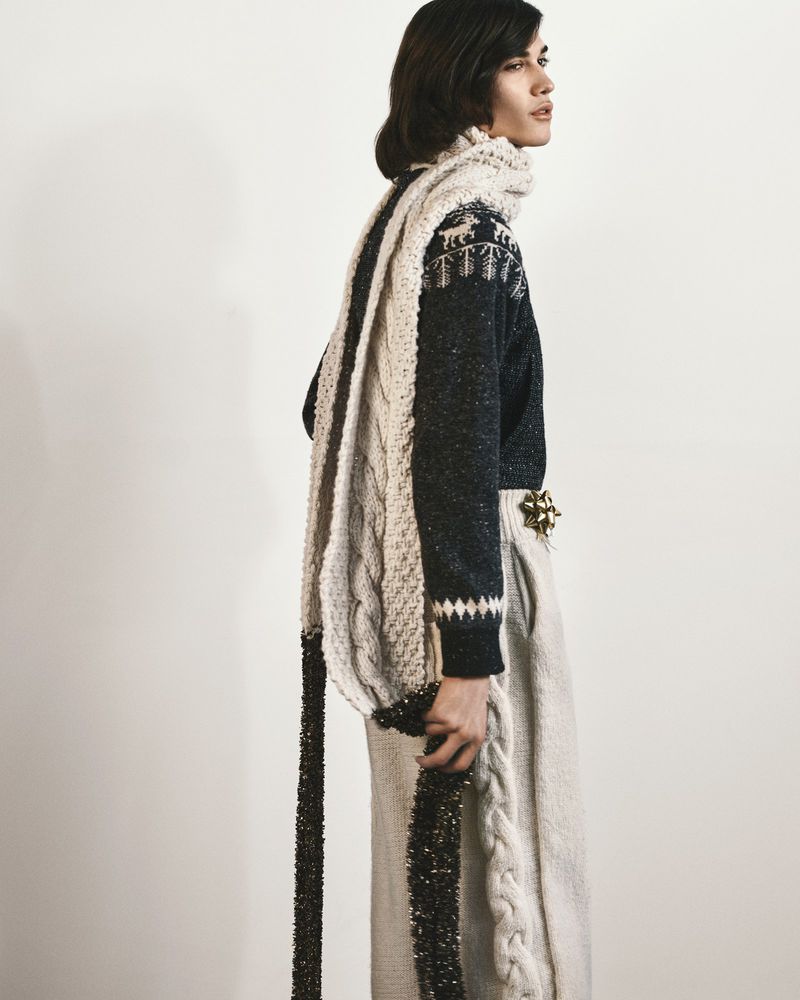 Model in profile wearing a dark patterned knit sweater and draped cream cable-knit scarf against a white studio backdrop.