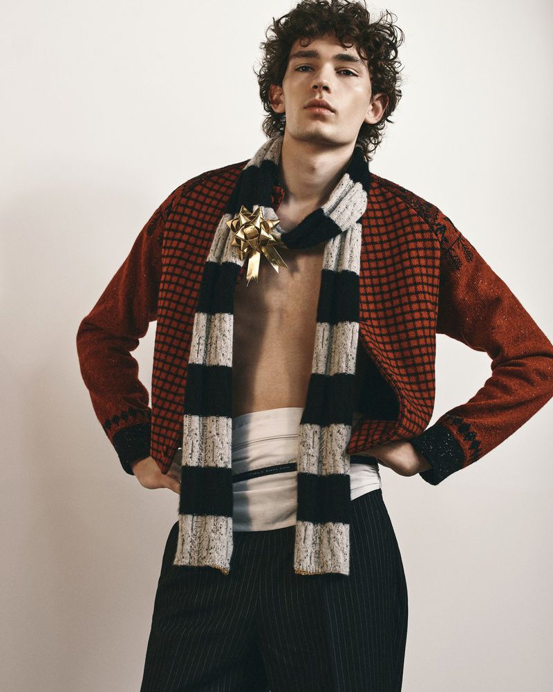 Model wearing an open red patterned cardigan, striped scarf with a gold bow, white sash, and pinstripe trousers against a white studio wall.