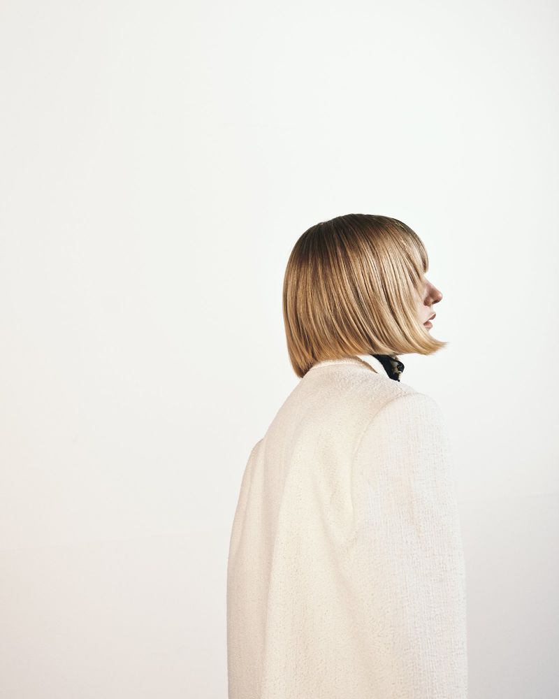 Model with a sleek blonde bob hairstyle viewed in profile wearing a cream textured blazer against a plain white studio backdrop.