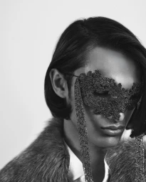 Monochrome close-up of a person wearing an ornate textured lace masquerade mask, fur coat, and white collared shirt.