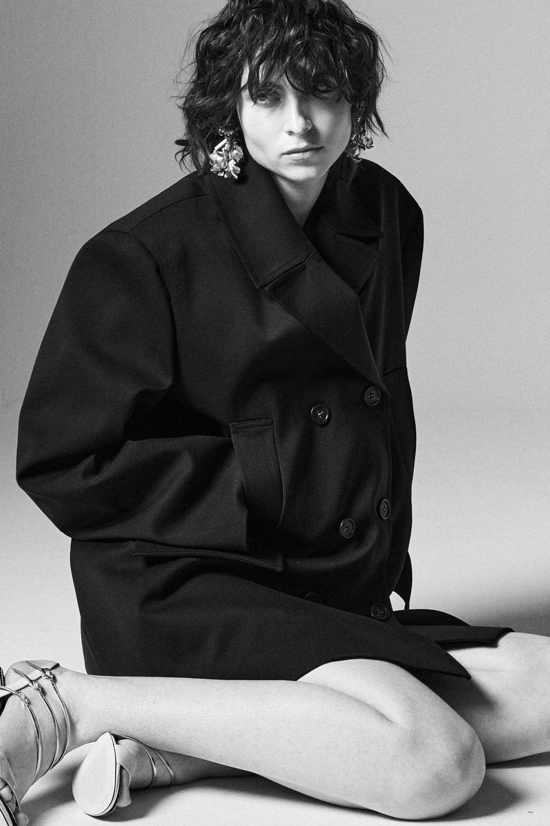 Model Evie Nopere seated in an oversized black double-breasted coat and strappy heeled sandals against a studio backdrop.