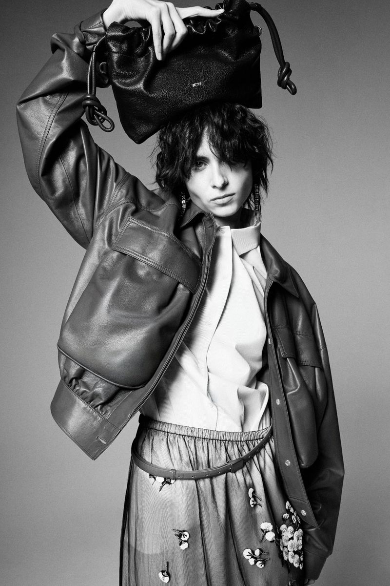 Evie Nopere holding a black leather pouch bag above her head, wearing an oversized leather jacket and sheer skirt in studio.