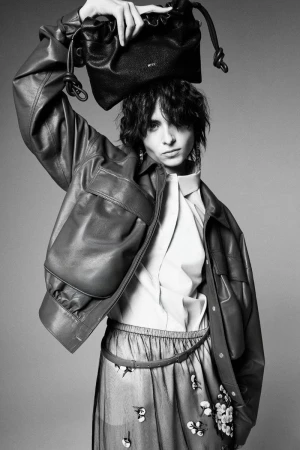 Evie Nopere holding a black leather pouch bag above her head, wearing an oversized leather jacket and sheer skirt in studio.