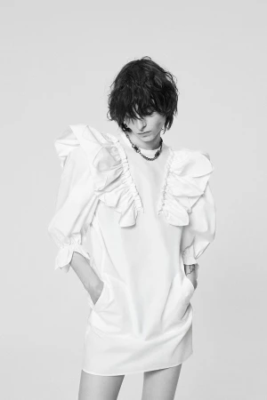 Model wearing a white ruffled dress with hands in pockets against a plain grey backdrop in N21 Resort 27 project.