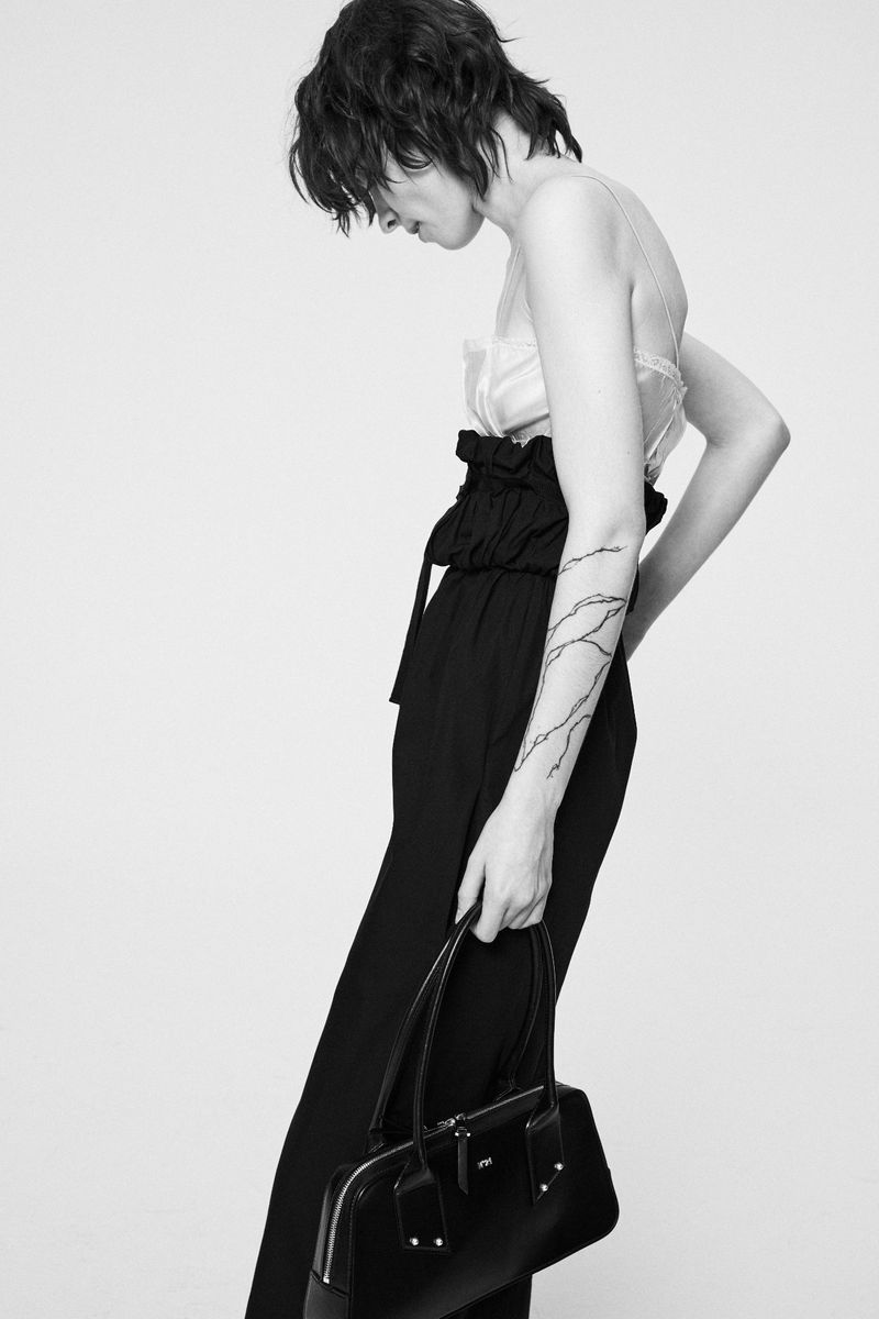 Model with shaggy hair in high-waisted black trousers holding a structured black leather handbag against a plain white studio background.