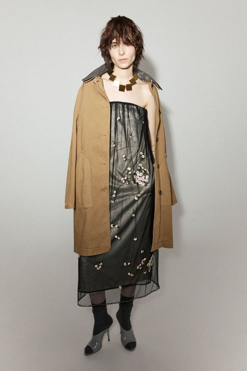 Model Evie Nopere poses in a camel coat, sheer floral dress, and chunky gold necklace against a grey seamless background.