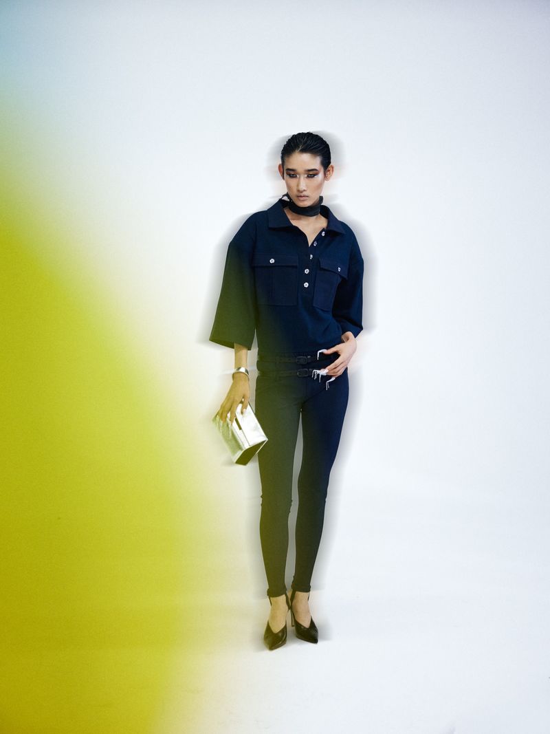 Model posing in a navy collared shirt, black skinny trousers, heels, and holding a metallic silver clutch against a studio backdrop.