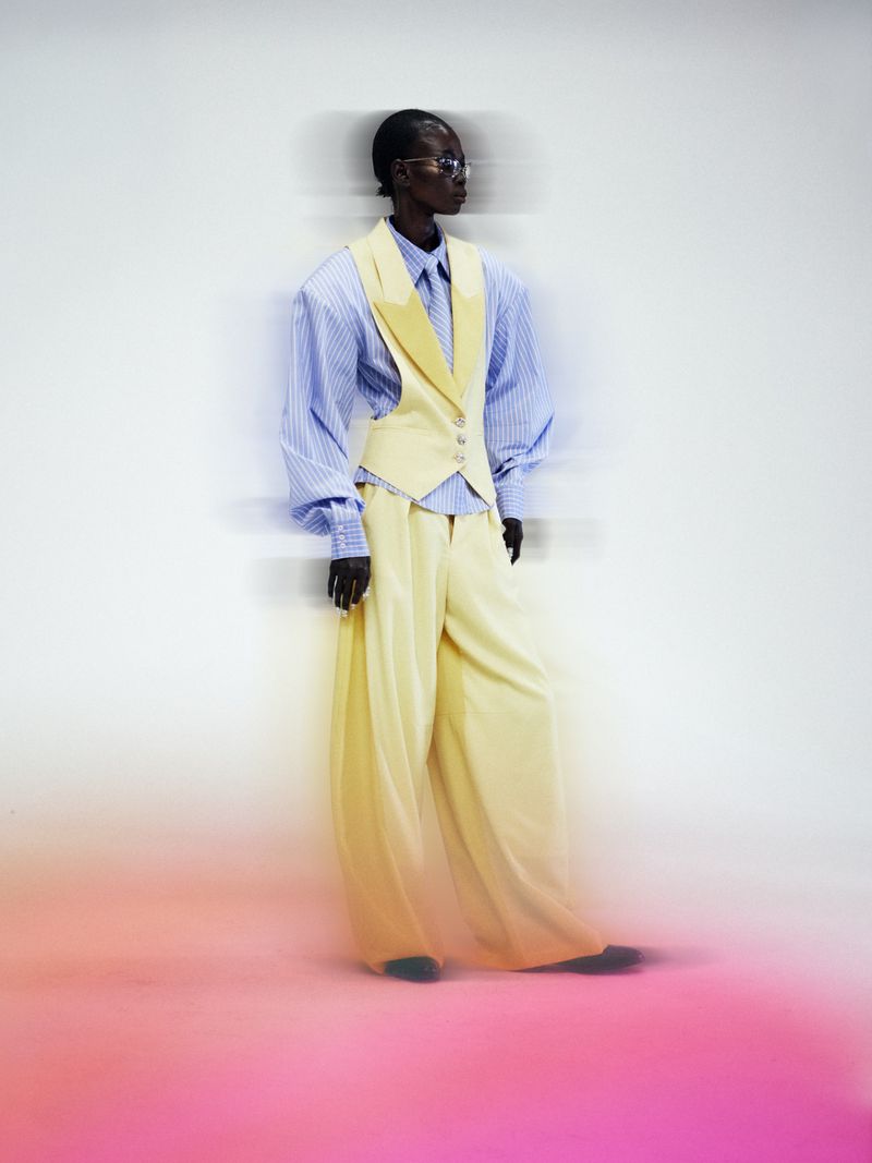 Model wearing a pale yellow waistcoat, matching wide-leg trousers, and a blue striped shirt against a white and pink studio backdrop.