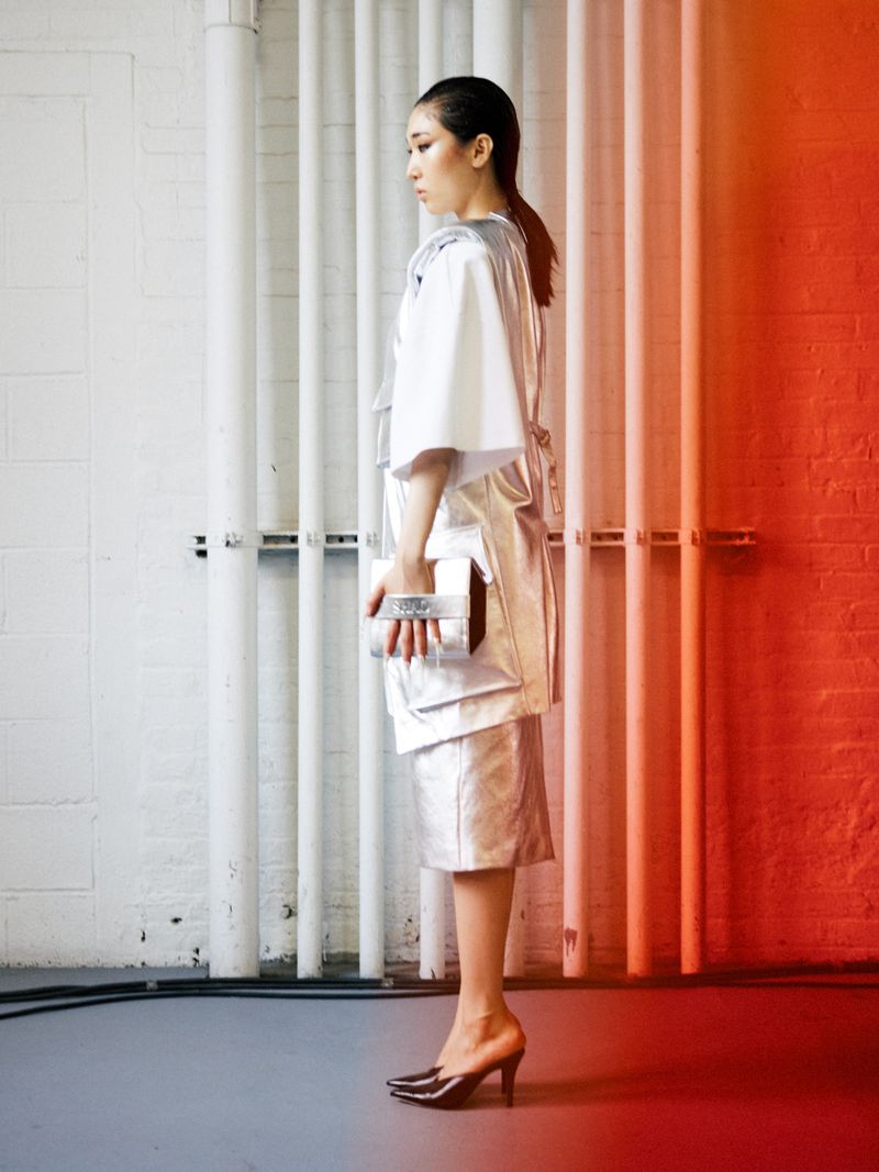 Model in profile wearing a metallic silver tunic, skirt, and white blouse holding a silver clutch against industrial pipes.