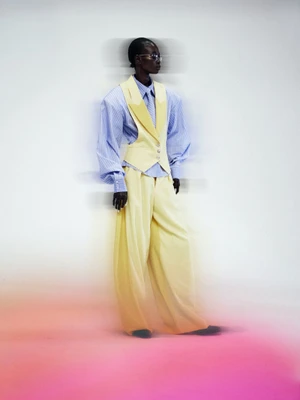 Model wearing a pale yellow waistcoat, matching wide-leg trousers, and a blue striped shirt against a white and pink studio backdrop.