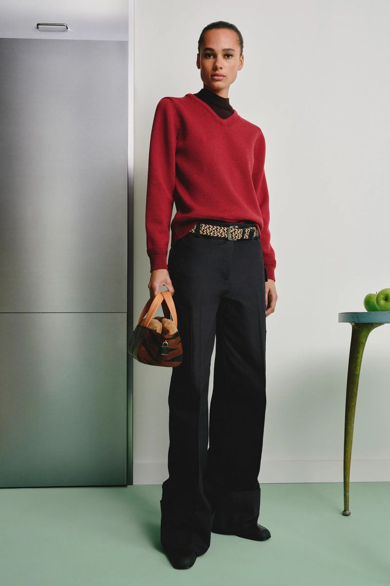 Claudia Campana wearing a red v-neck sweater, black trousers, and a patterned belt holding a handbag for Tory Burch.