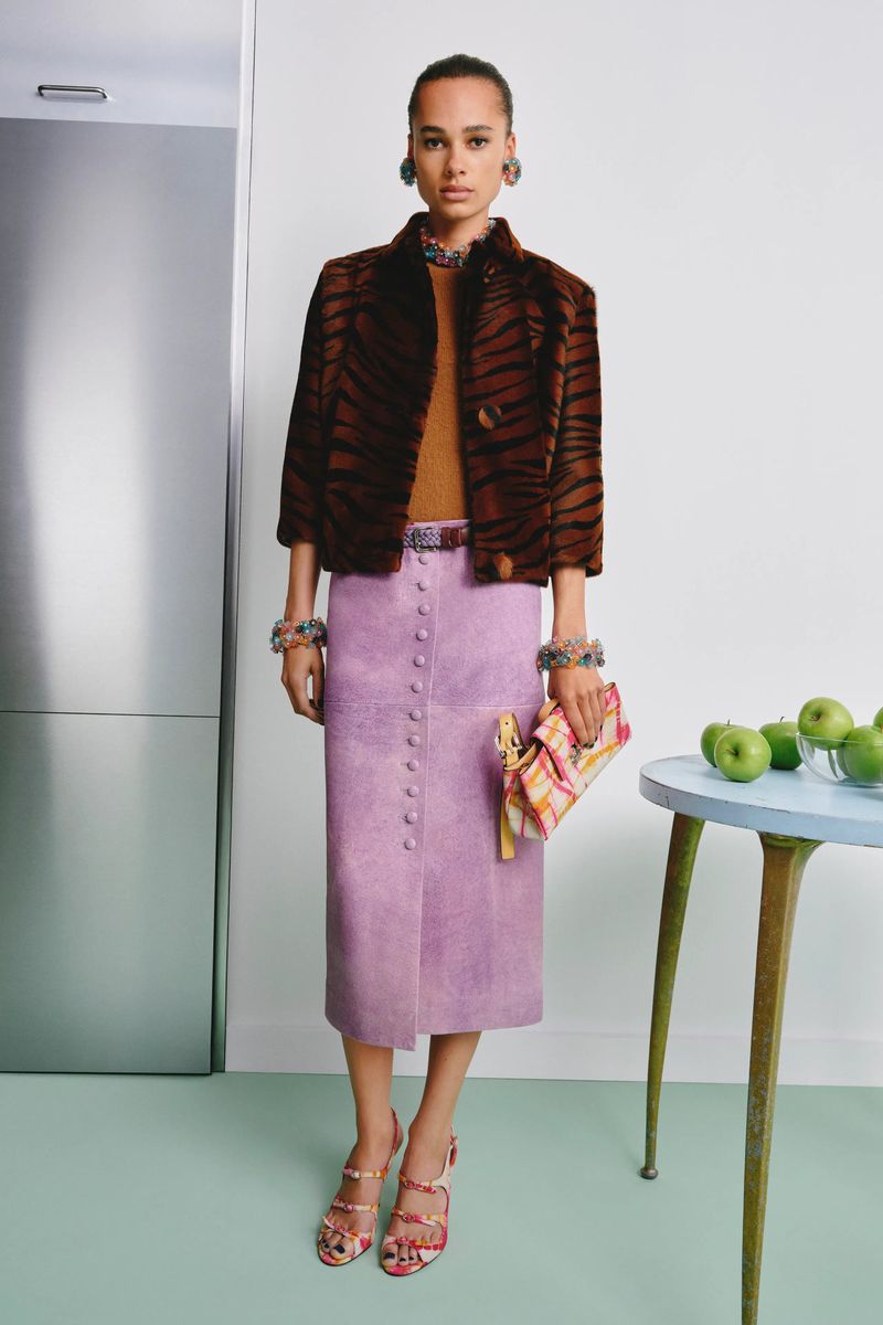 Model standing in a brown tiger-stripe jacket and lilac skirt holding a patterned handbag in a studio set.