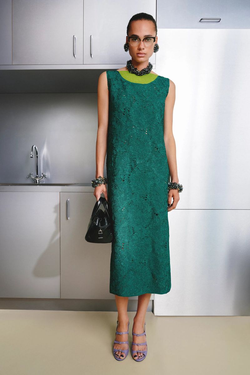 Model wearing a green textured dress and black handbag standing in a stainless steel kitchen interior