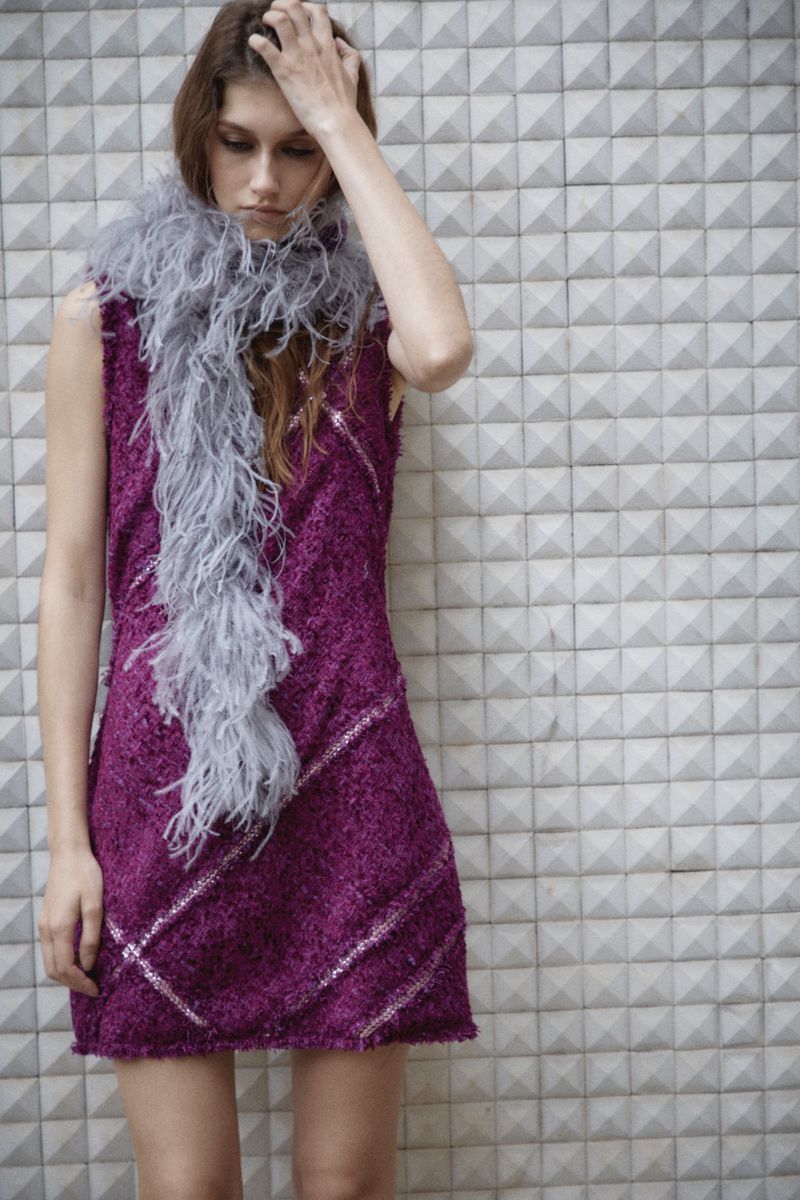 A young woman poses against a geometric wall wearing a purple dress with a grey feathered scarf, creating a fashionable and edgy look.