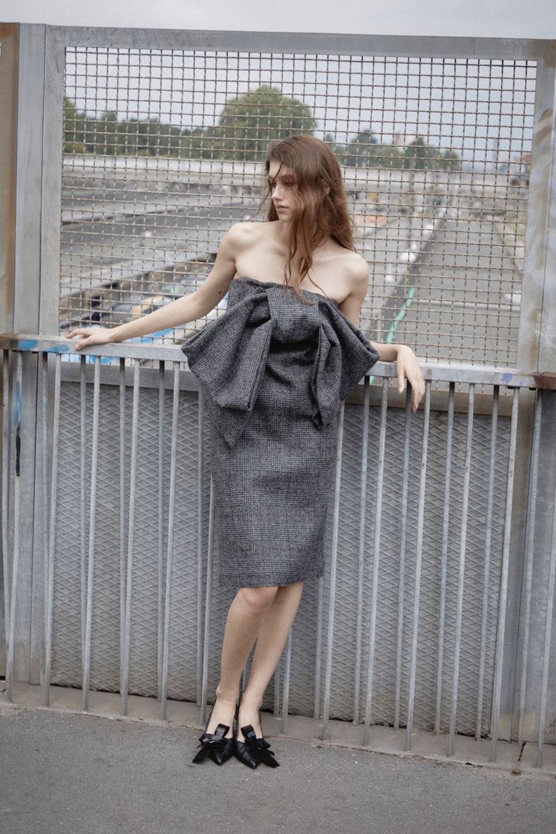 A woman poses in a strapless grey dress with a large bow, standing on a bridge with a metal fence and industrial background.