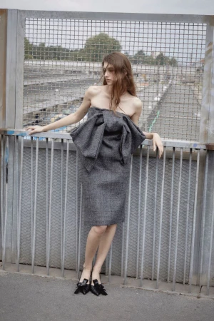 A woman poses in a strapless grey dress with a large bow, standing on a bridge with a metal fence and industrial background.