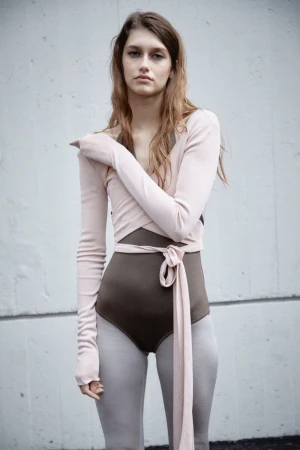 A young woman poses in a beige long-sleeve wrap top and brown bodysuit, set against a neutral wall, in a studio fashion shot.