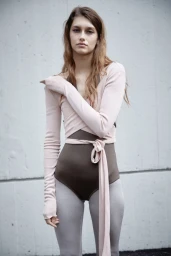 Model Roxane Marie in a pink wrap top, brown leotard, and grey tights standing against a concrete wall for EN POINTE.