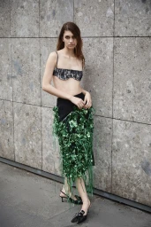 Model Roxane Marie posing against a textured stone wall in a cropped bralette, green metallic fringe skirt, and black slingback heels.
