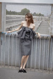 Model in a strapless grey tweed dress with a large bow detail leaning against a metal railing outdoors.