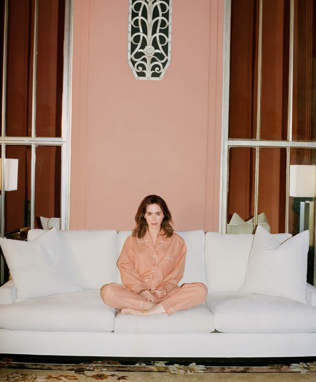Emily Blunt seated cross-legged on a white sofa in peach pyjamas against a pink wall for WSJ