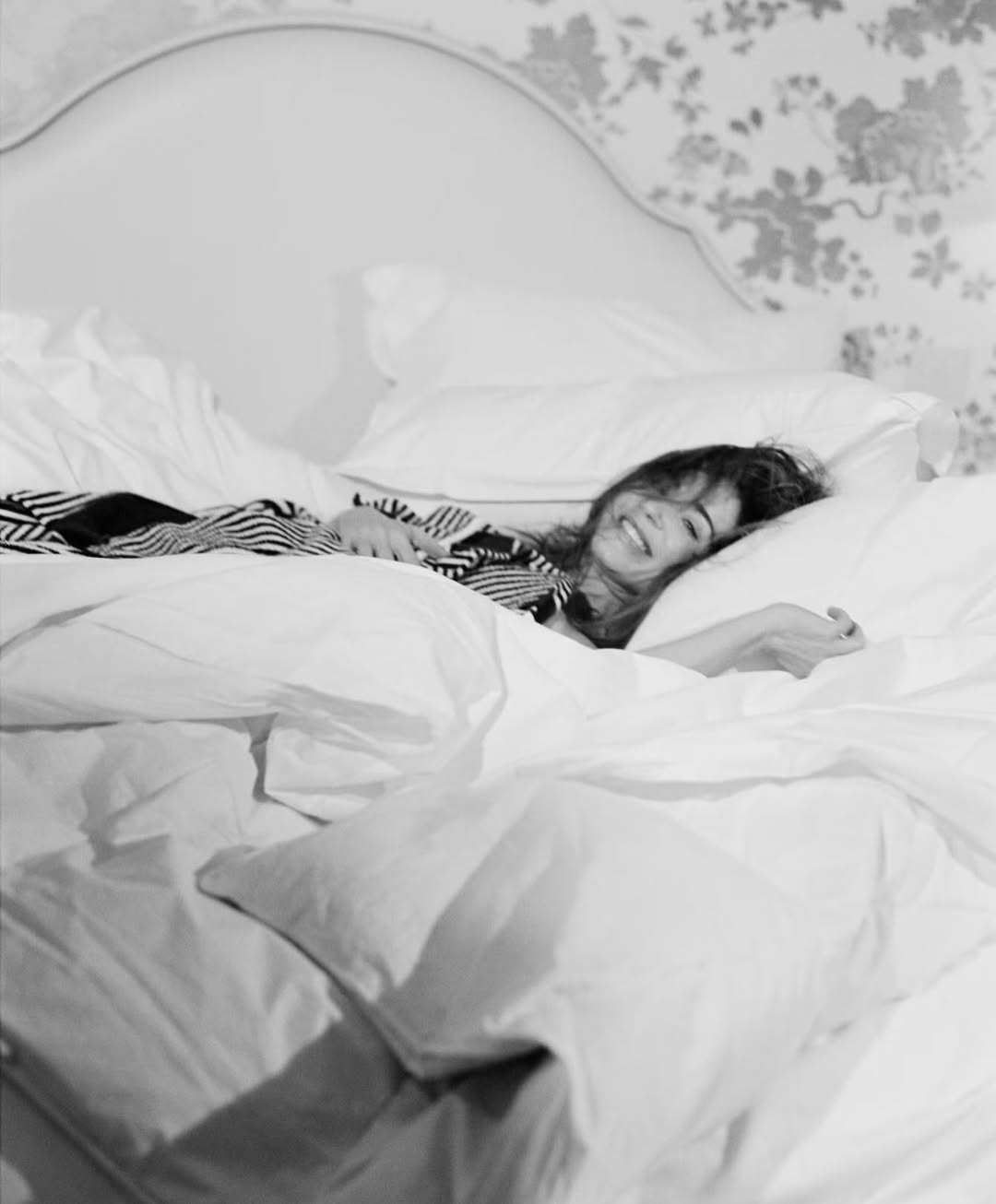 Emily Blunt lying in a bed with white sheets and pillows, smiling in a black and white editorial portrait.