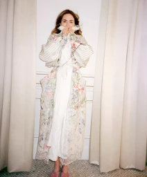 Emily Blunt standing between cream drapes wearing a voluminous floral embroidered white coat over her face.