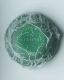 A round green skincare bar wrapped in crinkled translucent paper with Gezeiten text visible in the center against a white backdrop.