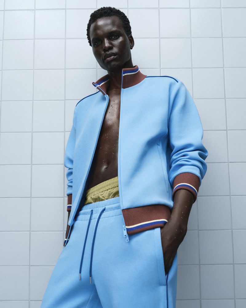 Model Badhiel Lony Nyang wearing an unzipped sky blue tracksuit against a white tiled wall for Rains.