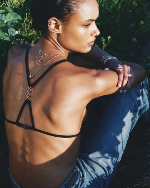A woman with dark hair and tanned skin poses outdoors wearing a black bra and blue jeans, accessorized with multiple necklaces and earrings.