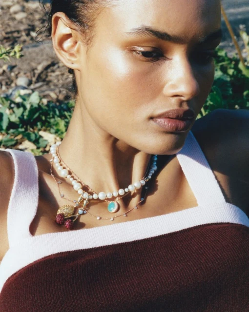 Close-up of a woman outdoors wearing a burgundy tank top and layered pearl necklace with cherry and pendant details.