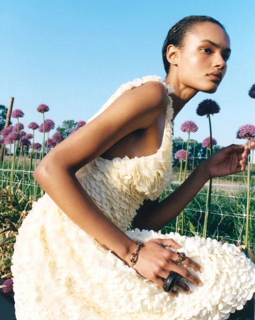Woman in ruffled yellow dress posing with purple allium flowers in an outdoor garden setting under a clear blue sky. Fashion editorial.