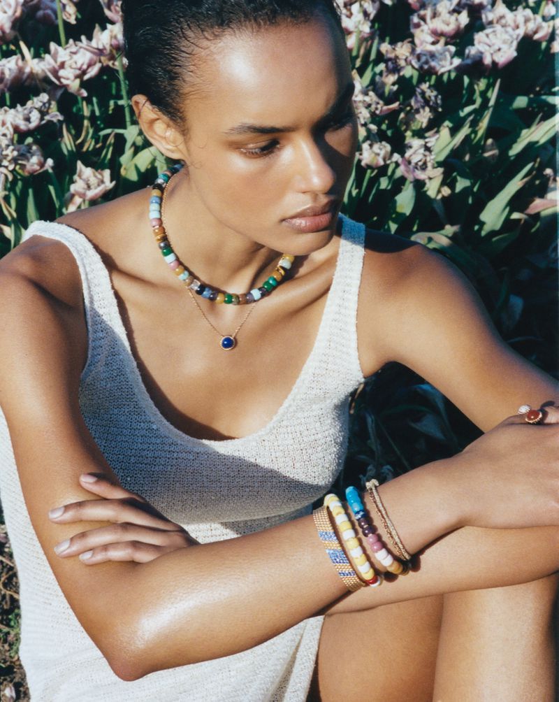 Model Ana Beatriz Cortes seated outdoors in a white knit dress wearing colorful beaded necklaces and stacked bracelets.