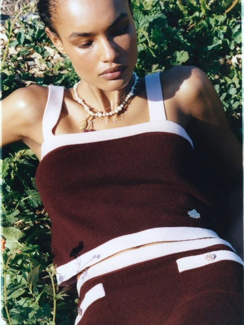 A brown-skinned woman with light brown hair, wearing a maroon and cream two-piece outfit, poses in a grassy outdoor setting.