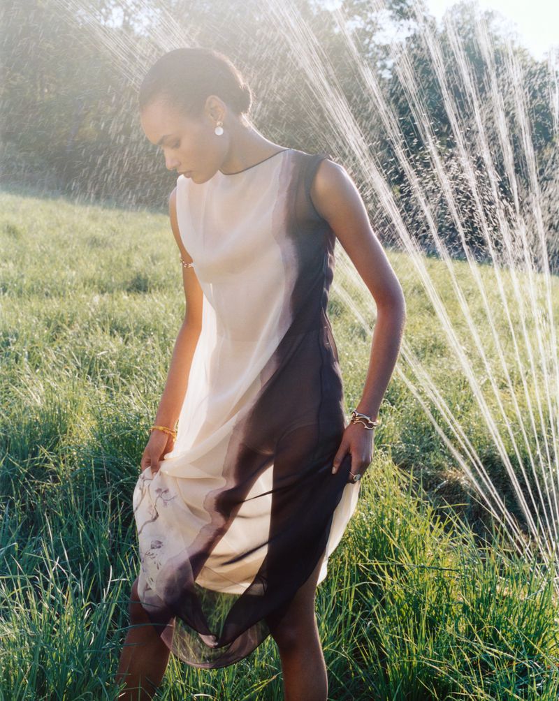 A young adult woman poses in a sheer dress in a grassy field with sunlight and a sprinkler system providing a soft, ethereal effect.
