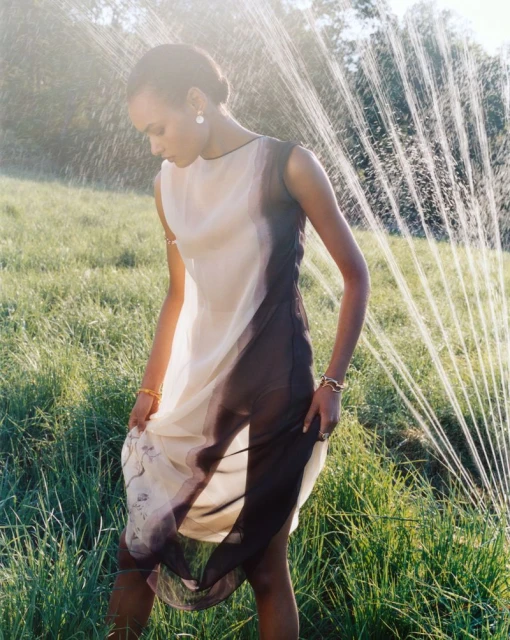 A young adult woman poses in a sheer dress in a grassy field with sunlight and a sprinkler system providing a soft, ethereal effect.