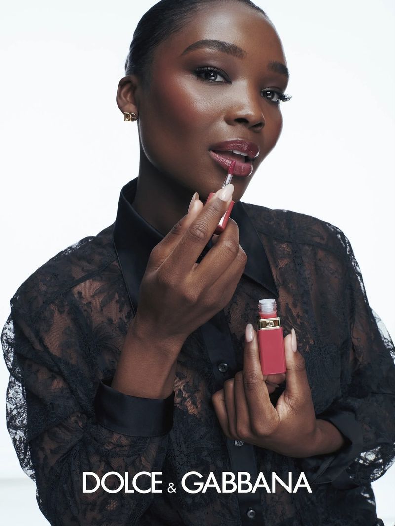 Melinda Melrose applying pink lip gloss while wearing a black sheer lace blouse against a bright white seamless background.